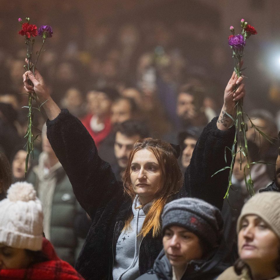 Antakya, Türkiye.  Amidst thousands of people, a woman holds up flowers at a rally on the anniversary of the earthquake.  A year ago, tens of thousands of people lost their lives in a massive earthquake in southeast Turkey.